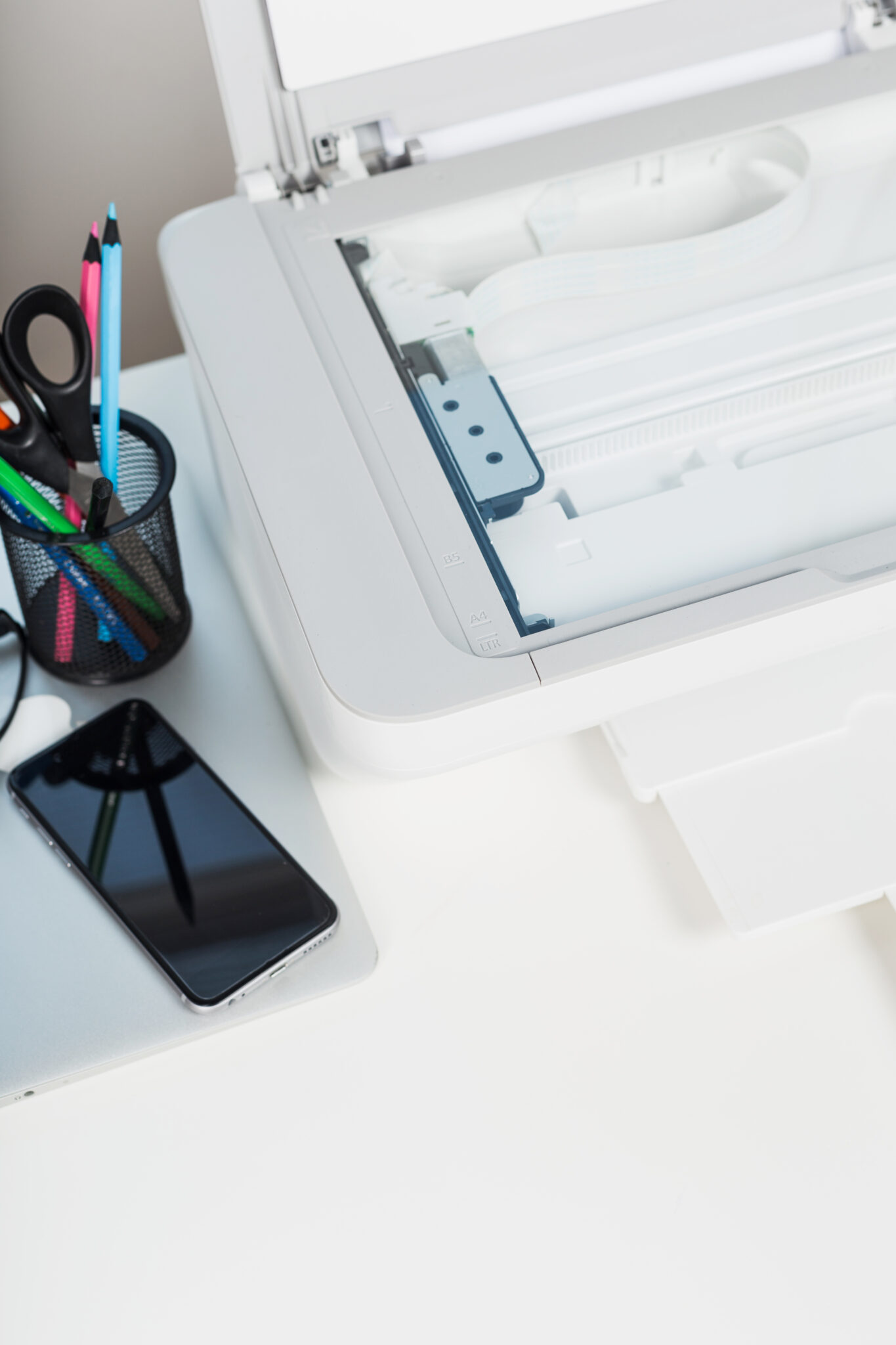 A modern workspace showcases a printer ready for use, accompanied by a smartphone and a holder filled with colored pencils. Bright daylight enhances the clean and organized setup.